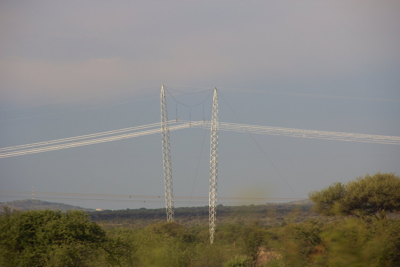 Steel electricity pylons and power lines across open ground, with low hills behind them.